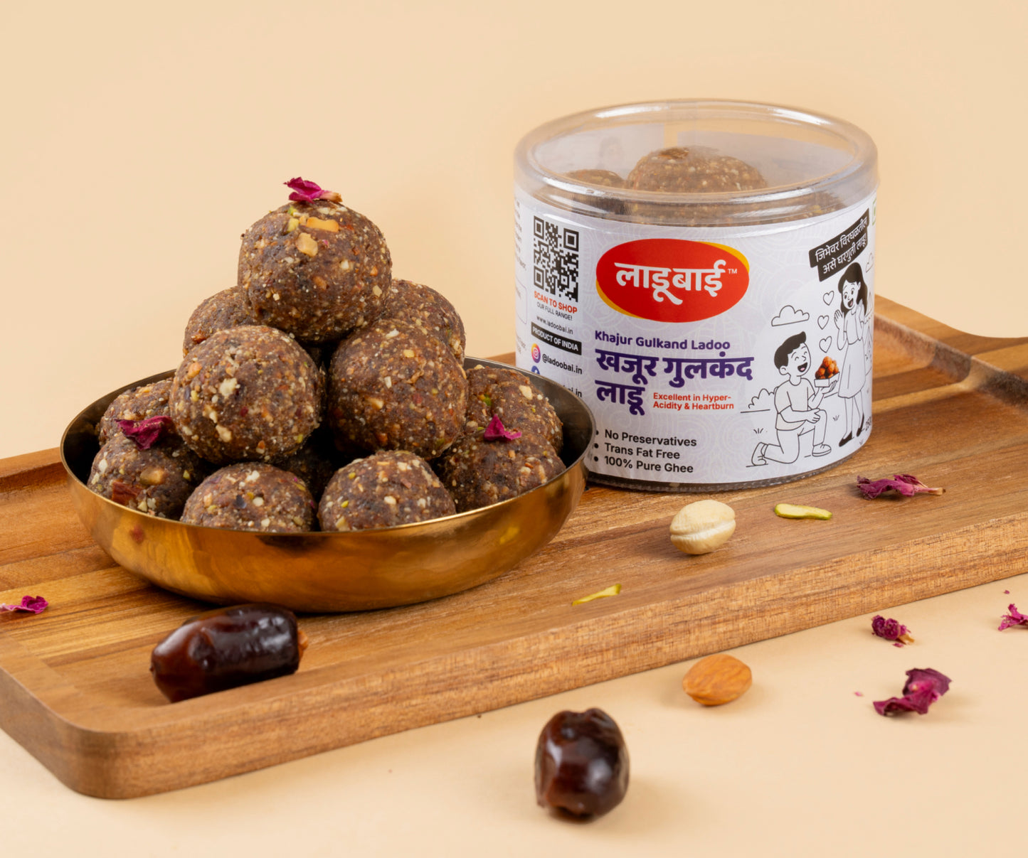 Khajur Gulkand Laddoos / Balls in a copper bowl with a container labeled 'Khajur Gulkand Ladoo' and Ladoobai's Brand and an illustration of a boy proposing a girl with laddoos, placed on a wooden board. Sprinkled ingredients/dry fruits around.