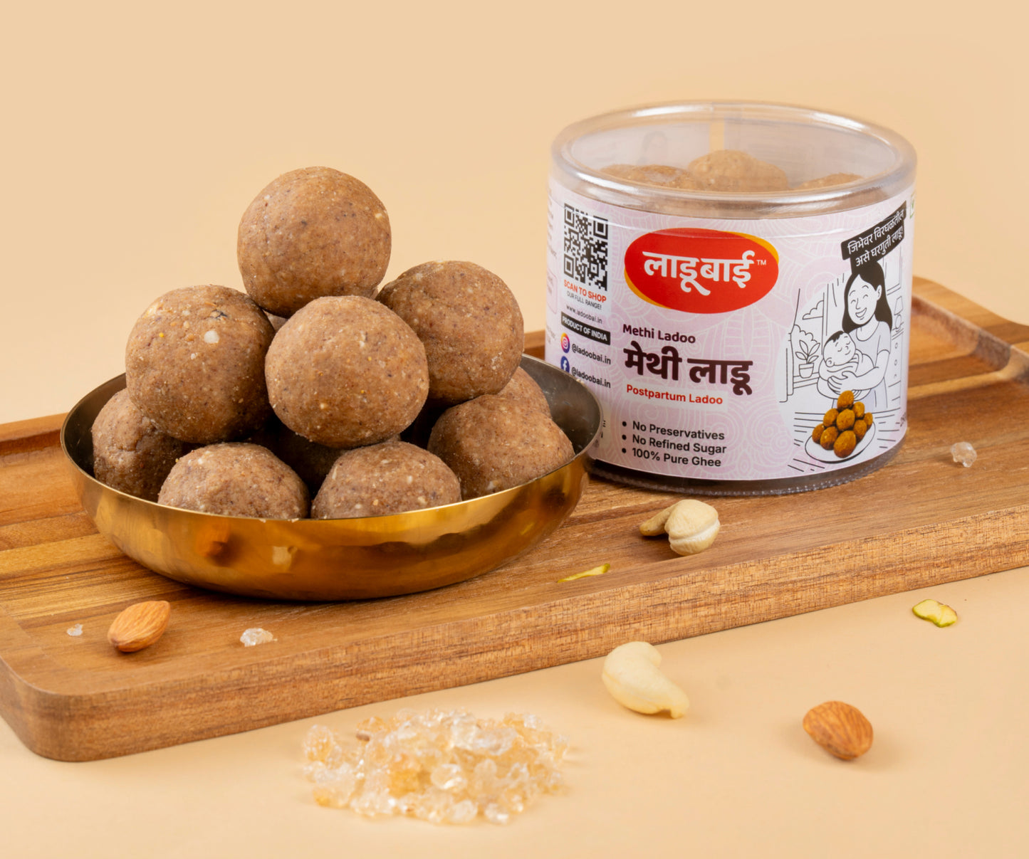 Light brownish Methi Laddoos / Balls in a copper bowl with a container labeled 'Methi Ladoo' and Ladoobai's Brand & illustration of a mother and a newborn child, placed on a wooden board. Sprinkled ingredients/dry fruits around.