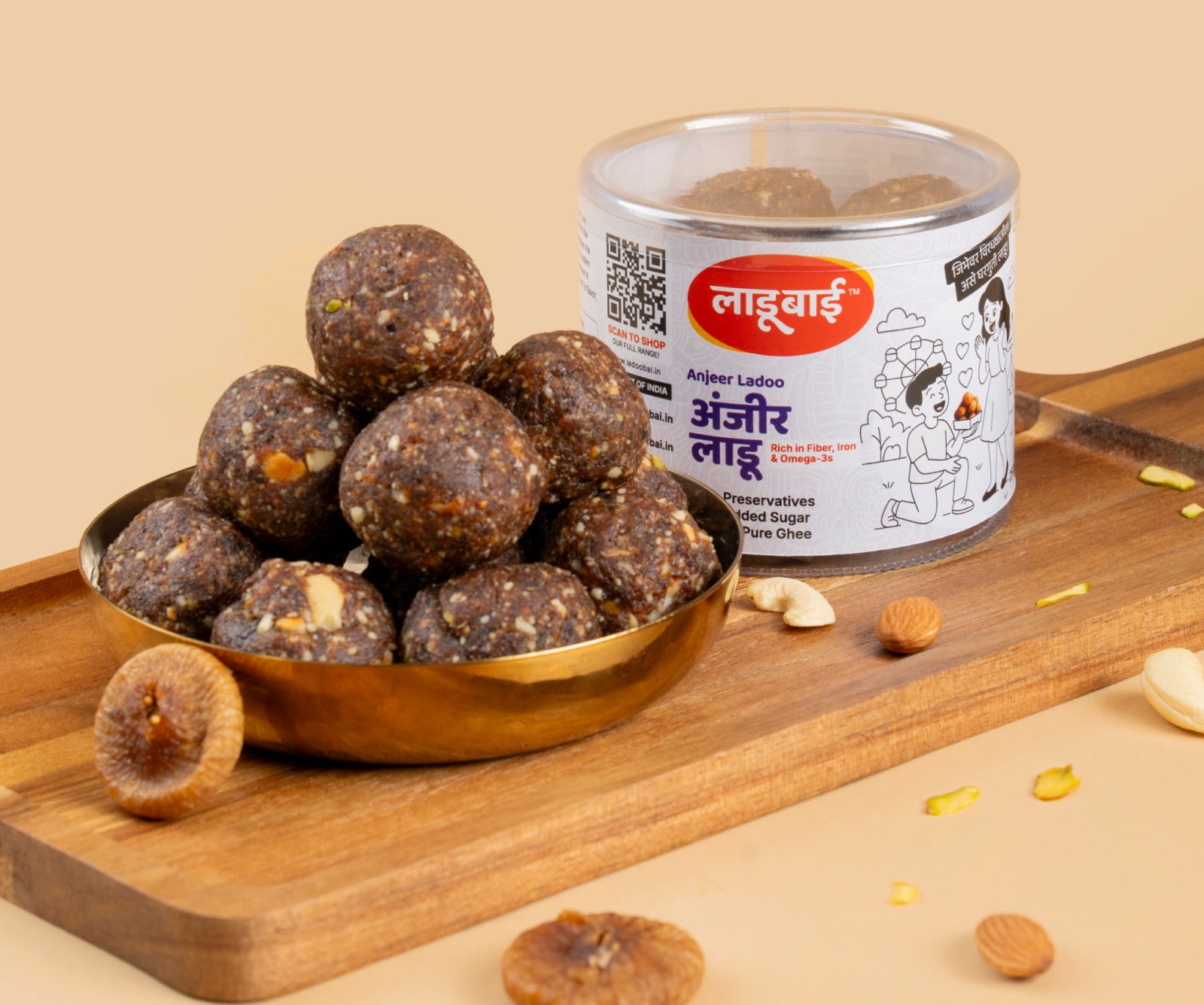 Anjeer/Dryfruit Laddoos / Balls in a copper bowl with a container labeled 'Anjeer Ladoo' and Ladoobai's Brand, placed on a wooden board. Sprinkled ingredients/dry fruits around.