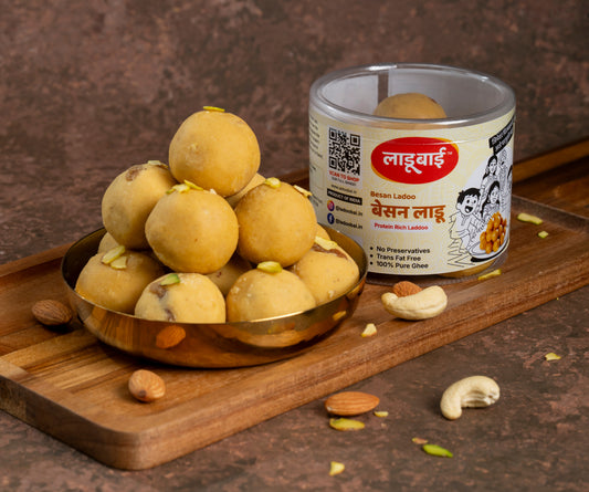Yellow golden Besan Ladoo / Balls in a copper bowl with a container labeled 'Besan Ladoo' and Ladoobai's Brand with text written Protein Rich Ladoo, placed on a wooden board. Sprinkled dry fruits around.