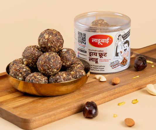 Dryfruit Laddoos / Balls in a copper bowl with a container labeled 'Dry Fruit Ladoo' and Ladoobai's Brand with text guilt free power snack, placed on a wooden board. Sprinkled ingredients/dry fruits around.