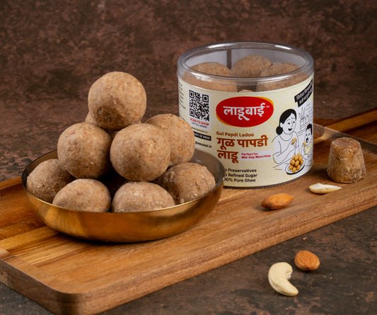 Light brownish Gul Papdi Laddoos / Balls in a copper bowl with a container labeled 'Gul Papdi Ladoo' and Ladoobai's Brand, placed on a wooden board. Sprinkled ingredients/dry fruits around and a small jaggery piece on the side.