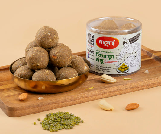 Light greenish Green Moong Laddoos / Balls in a copper bowl with a container labeled 'Hirva Moong Ladoo' and Ladoobai's Brand, placed on a wooden board. Sprinkled ingredients/dry fruits around and whole green moong bean on the side.