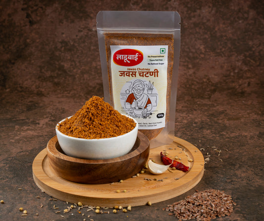 Spicy flax seeds chutney packet labelled 'Jawas Chutney' in Marathi language with ladoobai brand logo and old lady illustration on it, with a bowl of coursely grounded flax seeds chutney, placed on a circular wooden board, flax seeds and ingredients sprinkled around, against a brown background.