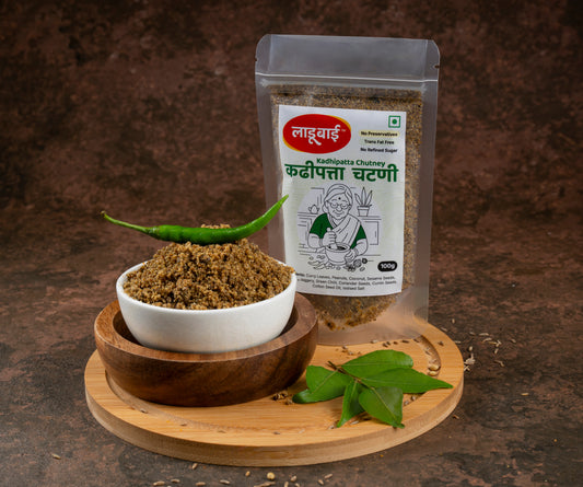 Spicy kadhipatta chutney packet labelled 'Kadhipatta Chutney' in Marathi language with ladoobai brand logo and old lady illustration on it, with a bowl of coursely grounded curry leaves chutney and green chilli placed on the heap, placed on a circular wooden board, curryleaves and ingredients sprinkled around, against a brown background.