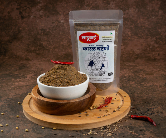 Spicy karal chutney packet labelled 'Karal Chutney' in Marathi language with ladoobai brand logo and old lady illustration on it, with a bowl of coursely grounded karal chutney, placed on a circular wooden board, ingredients sprinkled around, against a brown background.
