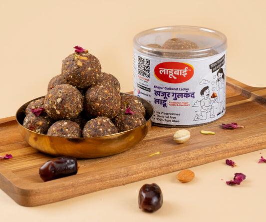 Khajur Gulkand Laddoos / Balls in a copper bowl with a container labeled 'Khajur Gulkand Ladoo' and Ladoobai's Brand and an illustration of a boy proposing a girl with laddoos, placed on a wooden board. Sprinkled ingredients/dry fruits around.