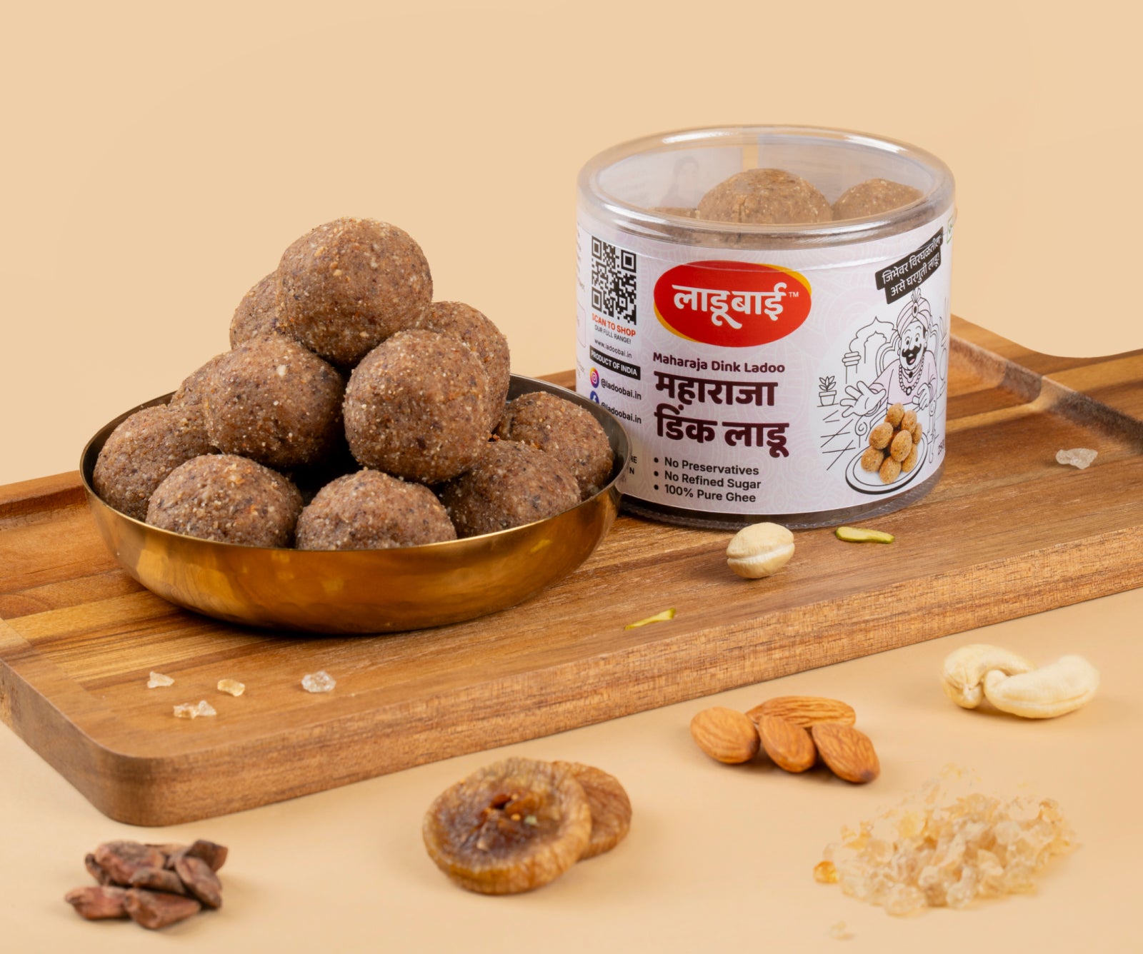 Light brownish Gond/Dink Laddoos / Balls in a copper bowl with a container labeled 'Maharaja Dink Ladoo' and Ladoobai's Brand and a king pleased reaching out to pick up a laddoo from a place, placed on a wooden board. Sprinkled ingredients/dry fruits around.