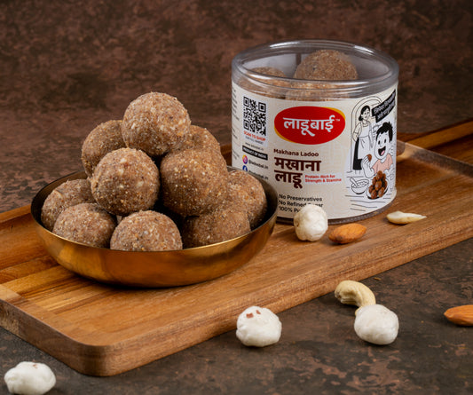 Round brown Makhana Laddoos / Balls in a copper bowl with a container labeled 'Makhana Ladoo' and Ladoobai's Brand, placed on a wooden board. Sprinkled ingredients/dry fruits around.