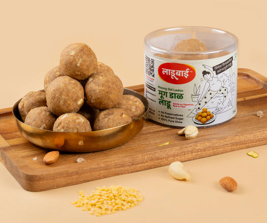 Light yellowish brownish Moong Dal Laddoos / Balls in a copper bowl with a container labeled 'Moong Dal Ladoo' and Ladoobai's Brand and illustration of a lady doing yoga and her cat, placed on a wooden board. Sprinkled ingredients/dry fruits around.