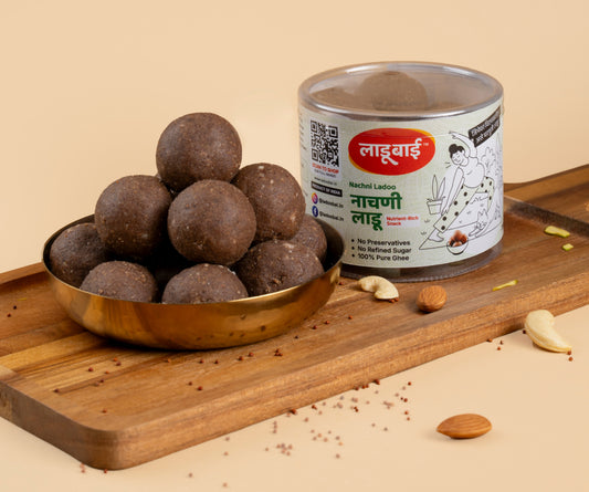 Dark brownish Nachni/Ragi Laddoos / Balls in a copper bowl with a container labeled 'Nachni Ladoo' and Ladoobai's Brand and illustration of a lady doing yoga and her cat, placed on a wooden board. Sprinkled ingredients/dry fruits around.