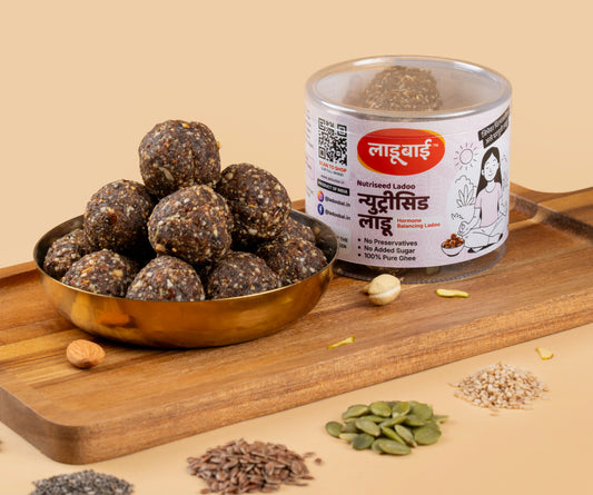 Brownish textured Nutriseed Laddoos / Balls in a copper bowl with a container labeled 'Nutriseed Ladoo' and Ladoobai's Brand and illustration of a lady meditating, placed on a wooden board. Sprinkled pumpkin seeds, til, flex seeds /dry fruits around.