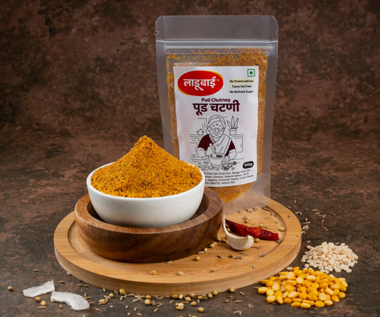 Spicy podi/pud chutney packet labelled 'Pud Chutney' in Marathi language with ladoobai brand logo and old lady illustration on it, with a bowl of coursely grounded lentil chutney, placed on a circular wooden board, lentils and ingredients sprinkled around, against a brown background.