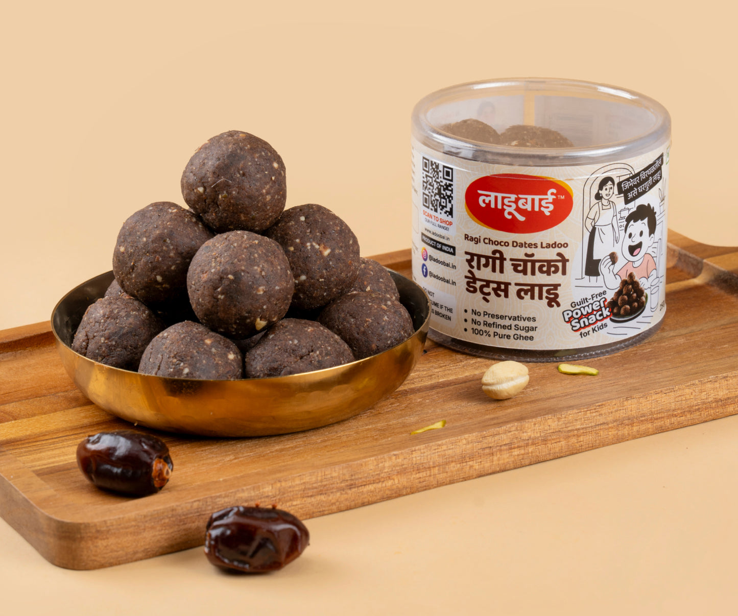 Dark brown Ragi Choco Dates Laddoos / Balls in a copper bowl with a container labeled 'Ragi Choco Dates Ladoo' and Ladoobai's Brand with text written as guilt-free power snack for kids, placed on a wooden board. Few dates & dry fruits spread around.
