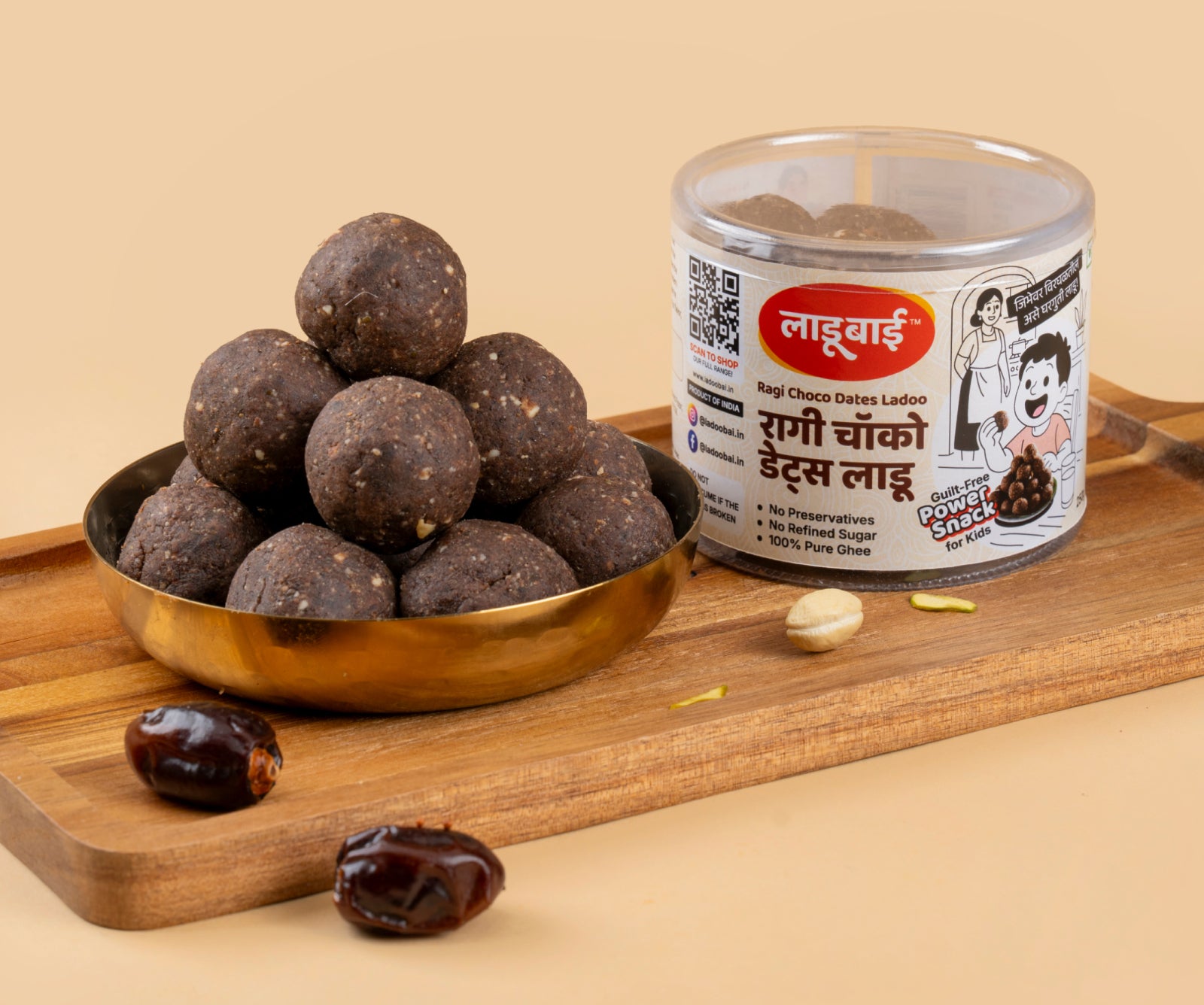 Dark brown Ragi Choco Dates Laddoos / Balls in a copper bowl with a container labeled 'Ragi Choco Dates Ladoo' and Ladoobai's Brand with text written as guilt-free power snack for kids, placed on a wooden board. Few dates & dry fruits spread around.