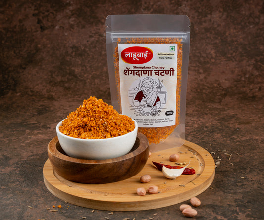 Spicy chutney packet labelled 'Shengdana Chutney' in Marathi language with ladoobai brand logo and old lady illustration on it, with a bowl of coursely grounded peanut chutney placed on a circular wooden board, ingredients sprinkled around, against a brown background.