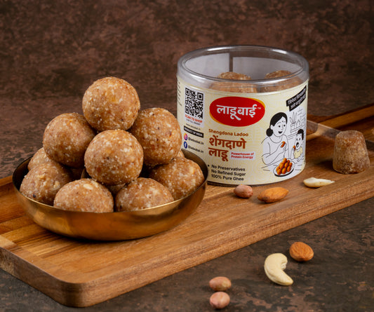 Light yellowish brownish textured Peanut Laddoos / Balls in a copper bowl with a container labeled 'Shengdana Ladoo' and Ladoobai's Brand and illustration of a lady and her kid enjoing laddoos sitting on a table placed on a wooden board. Sprinkled ingredients/dry fruits around and a jaggery piece on the side.