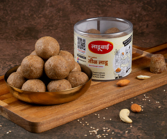Brownish textured Sesame/Til Laddoos / Balls in a copper bowl with a container labeled 'Til Ladoo' and Ladoobai's Brand and illustration of a old person reading paper sitting on a chair, placed on a wooden board. Sprinkled ingredients/dry fruits around and a jaggery cube on the side.