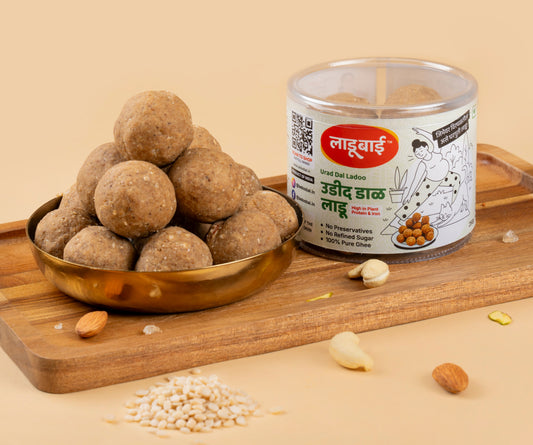 Light brownish Urad Dal Laddoos / Balls in a copper bowl with a container labeled 'Urad Dal Ladoo' and Ladoobai's Brand and illustration of a lady doing yoga and her cat, placed on a wooden board. Sprinkled ingredients/dry fruits around.